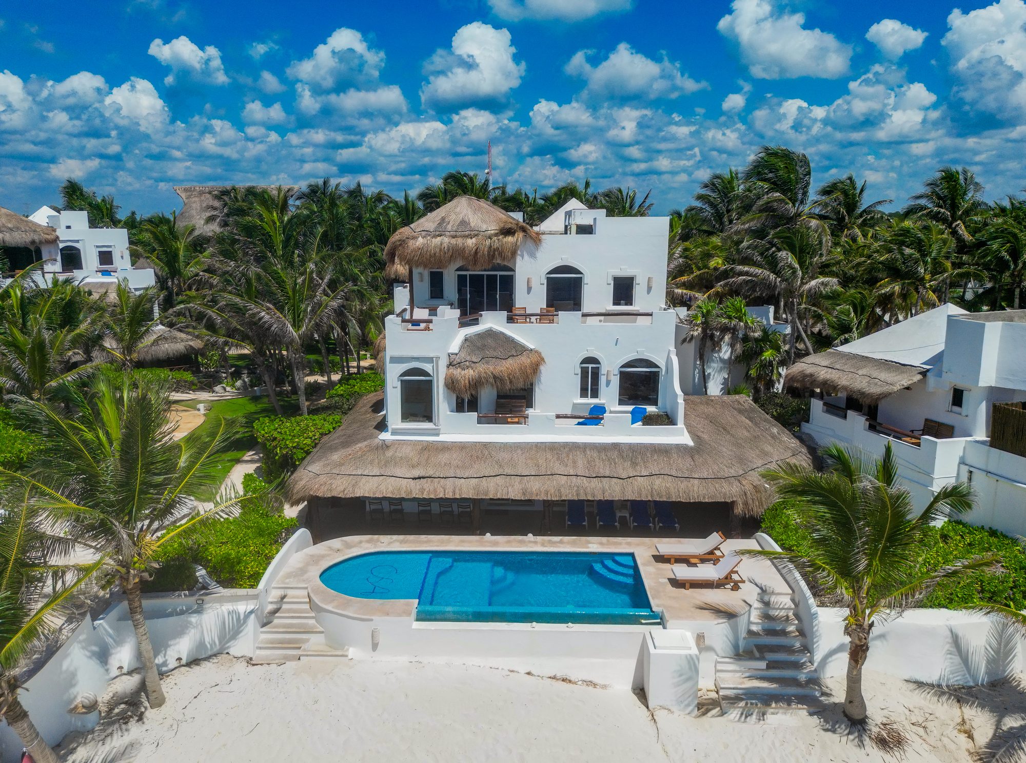 A three-story whitewashed estate villa under palapa roofs above its monogrammed beachfront pool