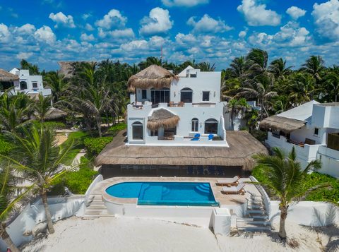 A three-story whitewashed estate villa under palapa roofs above its monogrammed beachfront pool