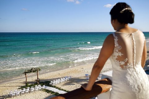 A bride on an estate balcony looking down at her beach ceremony set on the sand below