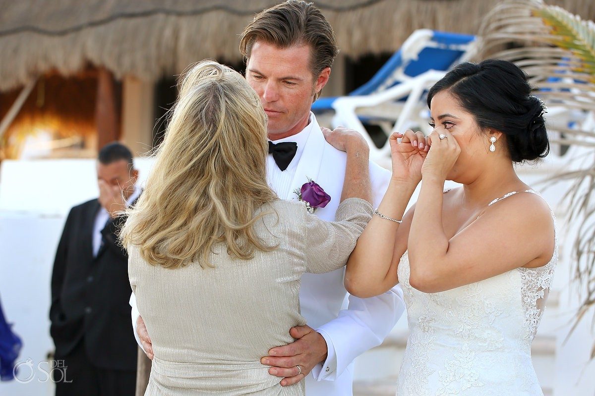 A bride wiping happy tears as the groom embraces his mother after the beach ceremony