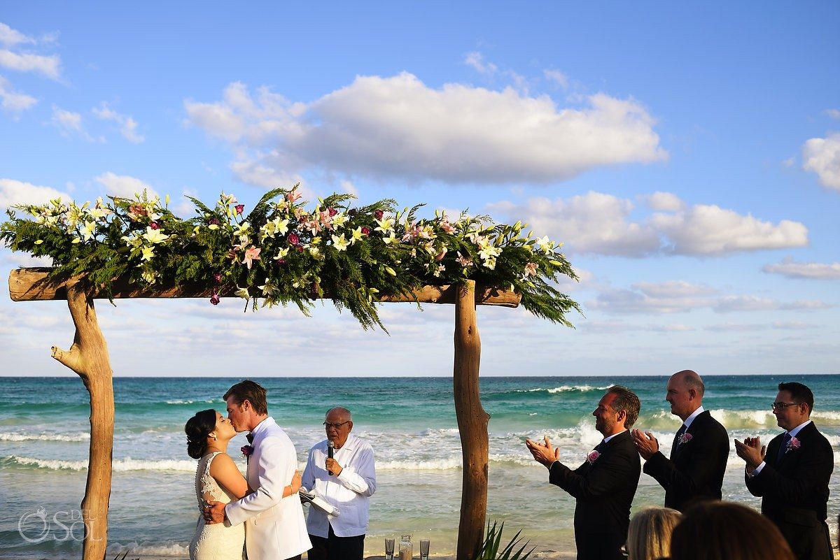 A couple's first kiss under a driftwood floral arch on the beach, the Caribbean behind them