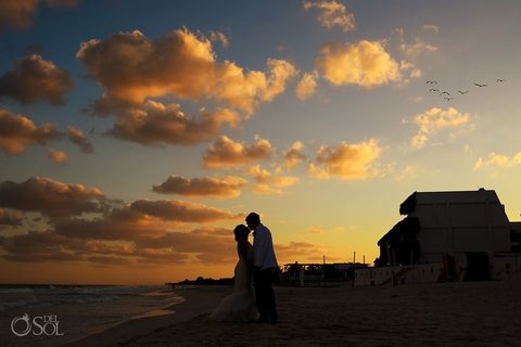 A couple silhouetted at sunset on the beach as pelicans fly past the estate's palapa